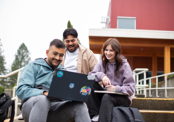 two men and a woman, all casually dressed, sit outside on stairs looking at a laptop. 