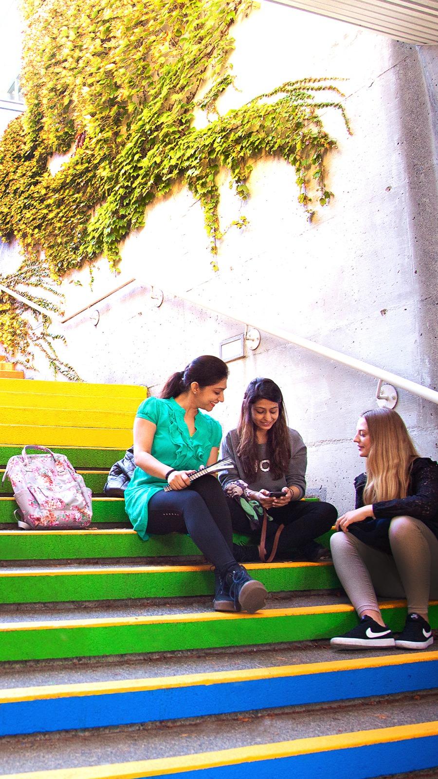 Three students sitting on colourful stairs with greenery above them. 