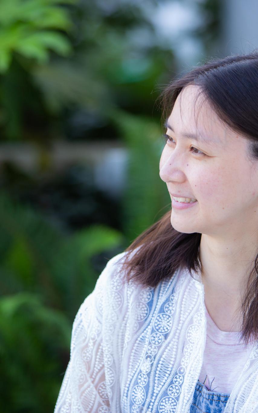 Female student with with shoulder length dark hair smiling and looking left in front of some trees.