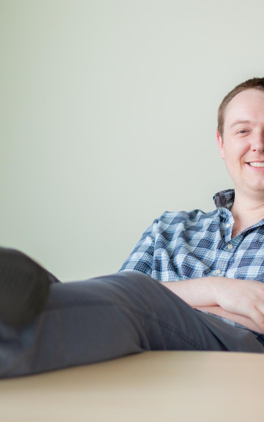 A man with short red-ish hair smiling in a blue plaid shirt with hid arms crossed and feet up on a table. 